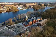 Luftbild eines länglichen, dreistöckigen Backsteinhauses mit Giebeldach und viereckigem Türmchen an der Seite umgeben von Bäumen, neben der Spree. Davor ein etwa halb so langer, dreistöckiger Anbau, und auf einer Freifläche daneben Bänke und Stühle zum Hinsetzen.