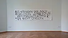 Auf einer weißen Wand hängt ein 1-Meter hohes und 3,2 Meter breites Papier. Es ist ebenfalls weiß bestrichen. Darauf steht in schwarzen, unsauberen, sich teils überlappenden Buchstaben: "AUS GRÜNDEN DER NACH HALTIGKEIT KANN EIN ÄS THETISCHER ANSPRUCH ALS ENTSTEHUNGSLEGITIMA TION NICHT GENÜGEN." Rechts auf dem Blatt ist ein sehr kleiner QR-Code aufgebracht, der zur Seite der Arbeit führt.