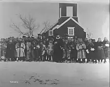 Schwarz-Weiß-Foto einer Gruppe von in Pelzmänteln gekleideter Personen vor einer Holzkirche