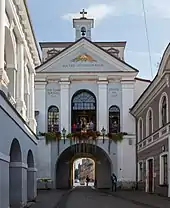 Frontale Farbfotografie eines blau-weißen Gebäudes mit einer Tordurchfahrt auf einer schmalen Straße. An beiden Straßenseiten stehen alte Gebäude. Über dem Tor ist eine Etage mit drei Rundbogenfenstern, in der Menschen stehen. Im darüber liegenden Giebelfeld ist ein goldenes Dreieck mit goldenen Strahlen und der lateinischen Aufschrift „Matea Misericordia“. Der hintere Dachbereich ist rechteckig und auf dem Giebel ist ein kleiner Glockenturm mit einem goldenen Jesuskreuz.