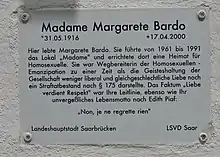 In schwarzer Schrift auf weißem Untergrund ist zu lesen: Madame Margarete Bardo *31.05.1916 +17.04.200 Hier lebte Margarete Bardo. Sie führte von 1961–1991 das Lokal „Madame“ und errichtete dort eine Heimat für Homosexuelle. Sie war Wegbereiterin der Homosexuellen. Emanzipation zu einer Zeit als die Geisteshaltung der Gesellschaft weniger liberal und gleichgeschlechtliche Liebe noch ein Strafbestand nach § 175 darstellte. Das Faktum „Liebe verdient Respekt“ war ihre Leitlinie, ebenso wie ihr unvergeßliches Lebensmotto nach Edith Piaf: Non, je ne regrette rein! Landeshauptstadt Saarbrücken LSVD Saar