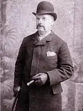Lusk hat einen Schnurrbart und trägt eine Melone, einen Mantel und Lederhandschuhe. In der rechten Hand hält er einen Stock, in der linken eine Zigarre.