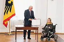 Frank-Walter Steinmeier zeigt die Urkunde, Gerda König lächelt. Daneben steht eine Deutschlandfahne.