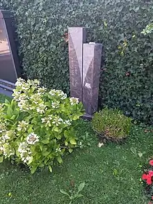 Grabstein von Hugo Loetscher: Hinter einem Hortensienbusch mit weissen Blüten in grüner Wiese und vor einer Efeuhecke stehen zwei quadratische Säulen unterschiedlicher Länge in rotem Marmorstein mit kleinstem Abstand auf einem Sockel nebeneinander. Schrift vertikal auf linker, längerer Säule: Hugo Loetscher, 1929–2009, auf rechter Säule aufgeschlagenes Buch mit Feder, seitlich Nummer: 81203 eingemeisselt. Friedhof Sihlfeld, Zürich.
