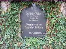 Schwarzer Grabstein mit goldener Schrift, an einer von Efeu bewachsenen Mauer angebracht. Die Inschrift lautet: Auguste Löber, geb. Feige, geb. 22. Nov. 1824, gest. 15. Jan. 1897, Begründerin der Auguste-Stiftung, „Selig sind die Bamherzigen, denn sie werden Bamherzigkeit erlangen.“, Matth. 5,7