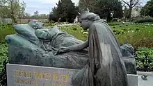  Fotografie. Im Vordergrund ist der Grabstein von Leopold Vander Kelen zu sehen. Der Grabstein ist eine Statue von zwei Personen. Sie zeigt den aufgebahrten Vander Kelen auf seinem Totenbett, die Arme vor der Brust verschränkt. Vor Vander Kelen kniet eine gekrönte Person, die einen Zweig neben den Toten legt. Der Sockel trägt die Inschrift „LEOPOLD VANDER KELEN“ und darunter „BOURGMESTRE DE LOUVAIN“. Im Hintergrund sieht man den Friedhof von Löwen.