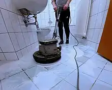 Eine Reinigungskraft reinigt mit einer Einscheibenmaschine einen Fliesenboden in einem Sanitärbereich