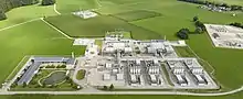Das Foto zeigt in der unteren Bildhälfte eine Industrieanlage inmitten grüner Felder. Links Parkplätze, dann ein Verwaltungsgebäude, ein Löschteich, ein Wendeplatz mit Waage für LKWs. Mittig folgt dann die Anlage selbst mit vielen Rohren, Rohrbrücken und eingehausten Anlagen. Etwas Abseits ein Materiallager und ein Umspannwerk. Rechts dann vier gleiche Anlagen mit je 4 Hochtanks. Links oben und am rechten Rand sind weitere eingezäunte dazugehörige Industrieanlagen.