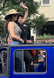 Hailie Sahar steht in einem dunkelblauen Auto mit offenem Dach und winkt der am Rand stehenden Menge zu. Sie trägt ein graues Top, eine Jeans, eine schwarze Sonnenbrille und einen beigenen Sonnenhut mit zwei gelben Streifen in der Mitte. Neben ihr steht ein Mann mit schwarzer Kappe, schwarzem T-Shirt und schwarzer Hose. Er hat einen regenbogenfarbenen Herz-Anstecker an der linken Brust und hält seine Hand auf Sahars Hüfte. Im Hintergrund sind ein Baum mit dunkelgrünen Blättern, ein beigenes Wohnhaus, ein Balkon mit spitzen, hellgrünen Pflanzen und Passanten verschiedenen Alters zu sehen.