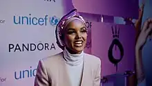 Halima Aden trägt ein rosafarbenes Kopftuch und cremefarbene Kleidung. Im Hintergrund ist eine Wand mit Werbung zu sehen.