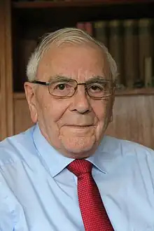 Porträt eines älteren Herrn mit grauen Haaren, Brille, hellblauem Hemd und roter Krawatte. Hintergrund in Unschärfe ein Bücherregal in Brauntönen