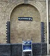 Frontale Farbfotografie von einer hellen Backsteinmauer mit dem Schild „Platform neundreiviertel“ in der Mitte. Ein Rundbogen umfasst das Schild und geht bis zum unteren, dunklen Mauerabschnitt. Ein Gepäckwagen steht in Fahrtrichtung an der Mauer.