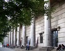 Die massive Fassade in diagonaler Ansicht, mit der Reihe der Säulen, der Freitreppe und einigen Ästen der Linden vor dem Gebäude