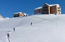 zwei große aus Holz erbaute Berghütte mit Schnee auf dem Dach auf einem Bergplateau, auf dem schneebedeckten Hang unter ihnen steigen Skiwanderer in einer S-Kurve auf