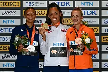 Anna Hall im blauem Trainingsanzug, Katarina Johnson-Thompson in weißer Trainingsjacke und Anouk Vetter in Orange präsentieren ihre Medaillen vor einer Sponsorentafel.