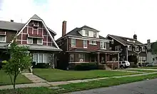 Blick von Straße auf drei stattliche zwei- bis dreigeschossige Wohngebäude. Alle mit Backsteinfassaden und dunklen Schindeldächern, das linke mit klaren Tudor-Elementen.