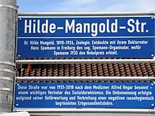 Foto des Straßenschilds in der Hilde-Mangold-Straße mit Erörterung darunter: Diese Straße war von 1931 bis 2018 nach dem Mediziner Alfred Hegar benannt – einem wichtigen Vertreter des Sozialdarwinismus. Die Umbenennung erfolgte aufgrund seiner Befürwortung und Verbreitung einer negativen rassehygienisch begründeten Erbgesundheitslehre.