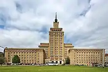 Frontale Farbfotografie eines mehrstöckigen, eckigen Turmgebäudes mit zwei langen Nebenbauten. Das mittlere Gebäude hat zwei Turmaufbauten und eine schwarze Spitze. Im unteren Bereich steht „Hotel International“. An beiden Seiten sind kleine Anbauten, vor denen die Nebenbauten stehen. Die Nebenbauten haben unter dem Dach rot-braune Ornamente.