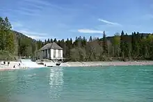 Über einem Wasserspiegel mit gletschergrünem Seewasser sieht man ein steiniges Ufer, das in Wald übergeht. In der linken Bildhälfte sieht man ein weißes Bauwerk mit hohen, schmalen Fenstern, neben dem sich schäumend Wasser über drei Stufen hinweg in den See ergießt.