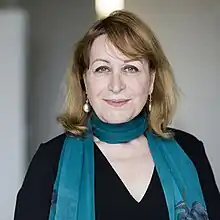 Das farbige Bild zeigt Kachachi in einer Naheinstellung vor einem unscharfen weiß-grauen Hintergrund. Kachachi lächelt den Betrachter an, hat halblanges dunkelblondes Haar und trägt zu einem schwarzen Oberteil einen blauen Schal, dessen Enden an ihrem Oberkörper hinabfallen.