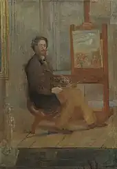 Ein Mann mit einem gezwirbelten Schnurr- und Kinnbart sitzt in aufrechter Pose vor einer Staffelei. In den Händen hält er Pinsel und Palette. Die Leinwand zeigt ein Jesusbildnis, bei dem der strahlende Heiligenschein auszumachen ist.