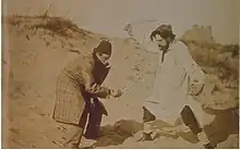 Sepia-Fotografie von zwei kostümierten Männern in einer Dünenlandschaft, die miteinander zu fechten scheinen. Der linke trägt einen karierten Mantel und einen Fez, der rechte einen hellen Kittel und eine Mütze. In den Händen halten sie Knochen und einen Totenschädel.