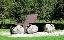 eine rostige Stahlplatte liegt auf großen Felsen, daraus ragt eine weitere rostige Stahlplatte schräg nach oben