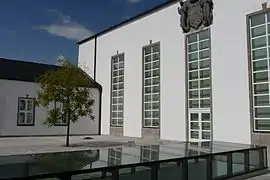 Links grüner Baum und kleineres weißes Gebäude, das an ein ebenfalls größes weißes links im rechten Winkel anschließt. Von diesem sind 4 hochgezogene rechteckige Fenster zu sehen. Über das dritte ist ein graues steinernes Wappen platziert. Auf dem gepflasterten Vorplatz ist teilweise ein nach oben hin verglastes niedrigereres Geschoss zu erkennen.