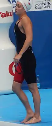 Emily Overholt trägt einen dunklen Badeanzug, bei dem ein Bein rot ist, und eine weiße Badekappe, auf der ein rotes Ahornblatt erkennbar ist und ihr Nachname in roter Schrift. In der linken Hand hält sie ihre Schwimmbrille. Im Hintergrund befindet sich eine blaue Wand mit Werbeaufschriften in lateinischer Schrift.