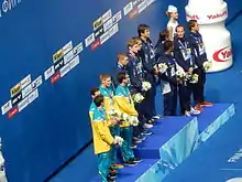 Foto von schräg vorne. Vorn die zweitplatzierten Australier in goldfarbener Trainingsjacke, dahinter die Sieger aus den Vereinigten Staaten und die Franzosen jeweils im dunkelblauen Trainingsanzug.