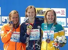 Die drei Schwimmerinnen posieren nach der Siegerehrung mit ihren Medaillen, die an einem blauen Band hängen. Die Sportlerinnen tragen ihre Trainingsjacke, an dem ihr Name auf Papier befestigt ist. Ashwood hält in ihrer linken Hand einen Blumenstrauß und das Maskottchen der WM.