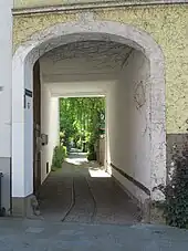 Torbogen in einer Hauserfassade mit geöffnetem Tor. Im gepfalsterten Boden sind Gleise verlegt, die vom Gehweg in Richtung Hinterhof wegführen