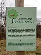 Text auf der Tafel: „Neuenrader Schnullerbaum.  Die Idee des Schnullerbaumes stammt aus Dänemark. Der Gedanke ist, dass das Kind die sonst eher problematische Trennung vom Schnuller mit einem positiven Erlebnis verbindet, da es den Schnullerbaum jederzeit besuchen kann. Gleichzeitig wird es auf diese Weise auch an die Natur herangeführt. Ebenso fällt der Verzicht auf den geliebten “Schnulli” leichter wenn es sieht, dass auch andere Kinder ihren Schnuller abgegeben haben. (Sic! Kommafehler nach „leichter“ im Original) Begleitet von Eltern, Großeltern, Tanten, Onkel und Freunden kann das Kind seinen Schnuller selbst an einen Ast hängen. Meist unterstützt ein kleines Geschenk der Eltern die Aktion und macht sie zu einem unvergessenen Ereignis. (Vom vorhergehenden Text abgesetzt und in anderer Farbe:) Achtung! Bitte verwenden Sie aus Naturschutzgründen nur neutrale Bindfäden zur Aufhängung. Keine Schleifenbänder, Kartons oder sonstiges Dekomaterial. Wir müssten sonst den Schnuller entfernen. forumneuenrade e. V. Abteilung Stadtverschönerung“