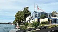 Zusehen ist das Ruderhaus mit den Bootshallen unten und dem Restaurant oben. Im Vordergrund sind die Stege zu sehen, über die die Boote zum See gelangen.