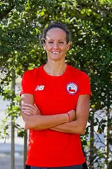 Kristel Köbrich steht im roten T-Shirt mit chilenischem Logo vor einem Baum. Die Arme hat sie vor ihrem Körper verschränkt.
