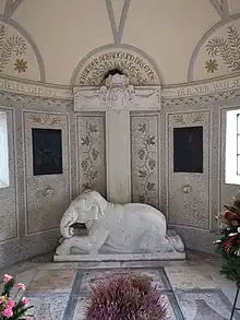Heller, runder Innenraum mit einem christlichen Kreuz unter dem ein Elefant liegt. Auf dem Kreuz ist ein Jesushaupt mit Dornenkrone zu sehen, darüber der Spruch "Ich bin der Anfang und das Ende"