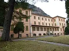 Das stillgelegte Kurhotel von Acquarossa ist von hohen Bäumen umgeben. Das imposante Gebäude hat zwei Seitenflügel und einen nach hinten gerückten Gebäudekorpus in der Mitte. Das Gebäude ist leicht rosa und die Fensterläden sind olivgrün. Alle Läden sind geschlossen. Der Garten und das Gebäude sind nicht heruntergekommen, aber ungepflegt.