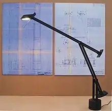 Schreibtischlampe Tizio. Auf einem zylindrischen Sockel ein dreigliedriges, bewegliches Gestell aus schwarzen Metallstreben, mit Gegengewichten ausbalanciert, am oberen Ende eine nach unten gerichtete Halogenlampe.