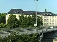 Eines der ur­sprüng­lichen Magazin­gebäude. Ansicht vom Nord­ost­rand des Lange­marck­platzes außer­halb der Liegen­schaft aus, Blick­richtung: süd­west, die auf dem Bild sicht­bare lange Seite des Ge­bäudes zeigt Rich­tung Nord­osten; die über die Brücke im Vorder­grund füh­rende Straße ist die B 416 Mayener Straße, das silberne Auto fährt Richtung Westen, unter der Brücke hindurch führt die B 9. (Die unten dar­gestellte Photo­post­karte zeigt dieses Ge­bäude vor Kriegs­be­schädigung und Wieder­aufbau).