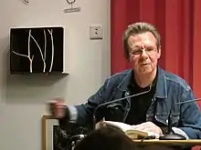 Siegfried Völlger an einem Tisch sitzend mit aufgeschlagenem Buch, spricht in ein Mikrofon
