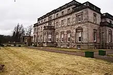 Whittingehame House. Das Bild zeigt einen Rasenfläche, dahinter das große Herrenhaus, ein dreistöckiges Gebäude mit jeweils 9 Fenstern zum Rasen hin, die sich nach britischer Bauweise nach oben hochschieben lassen. im Obersten Stockwerk sind je 7 Fenster verbaut, dazu gibt es eine Terrasse.