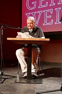 Ganzkörperbild eines Mannes mit kurzen grauen Haaren, der an einem Lesetisch sitzt und in ein Standmikrofon spricht. Er hält ein schmales weißes Buch aufgeschlagen in den Händen.