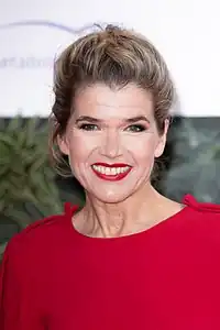 Ein Brustbild einer Frau in einem schlichten roten Kleid, die lächelnd in die Kamera schaut. Ihr Mund ist leicht geöffnet, so dass man ihre Zähne sieht. Ihre Lippen tragen roten Lippenstift. Die Haare sind hochgesteckt, der Hintergrund ist nur unscharf zu sehen.