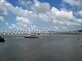 Die Brücke ist aus der Ferne zu sehen. Im Vordergrund ist Wasser und ein Boot. Der Himmel ist blau und leicht wolkig.