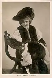 Madge Lessing ist sitzend auf einer schwarz-weiß Fotografie abgebildet. Sie trägt einen großen geschwungenen Hut, darunter ist helles Haar zu sehen. Sie trägt Kleidung aus Pelz.
