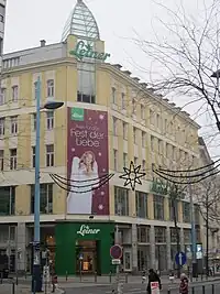 Foto eines Gebäudes an Straßenecke, optisch sehr ähnlich dem Foto 100 Jahre zuvor. An mehreren Stellen des Gebäudes prangt der Schriftzug "Leiner". Über dem Eingang hängt eine große Reklame mit der Aufschrift "Alles für das Fest der Liebe".