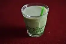 Ein transparentes Glas mit grün bedruckten Blättern beinhaltet eine hellgrüne Flüssigkeit. Es steckt ein dunkelgrüner Strohhalm im Glas, das auf dunkelrotem Untergrund steht, der das gesamte Bild ausfüllt.