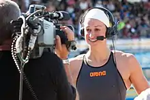 Megan Romano im dunklen Badeanzug mit Weißer Badekappe und darüber einem Headset wird gerade interviewt. Die linke Bildhälfte zeigt den Kameramann von hinten.