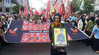 Porträt des YPG-„Märtyrers“ Şevket Demir (Çîya Amed) vor Transparent der „Suruç-Märtyrer“