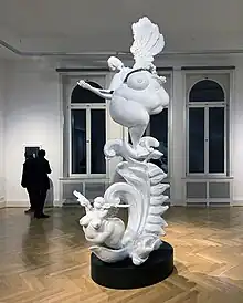 Auf einer barocken Ranke steht eine weibliche sehr üppige Frauenfigur. Die gedrehte und nach hinten abkippende Figur wirkt von weitem noch barock und üppig. Wer näherkommt und sie umrundet, erkennt, dass es sich um Fragmente einer Skulptur handelt, eine Hülle, eine Art Nachbild von Kraft und Vitalität. Aus ihrer abgesprengten Brust fliegt eine Nixe wie ein frisch geschlüpfter Schmetterling und auch am anderen Ende der Ranke schiebt sich eine Nixenartige Frauengestalt mit Flügeln aus einem Blatt. Vanitas und Neubeginn, Re-Naissance im Gefühls-Chaos